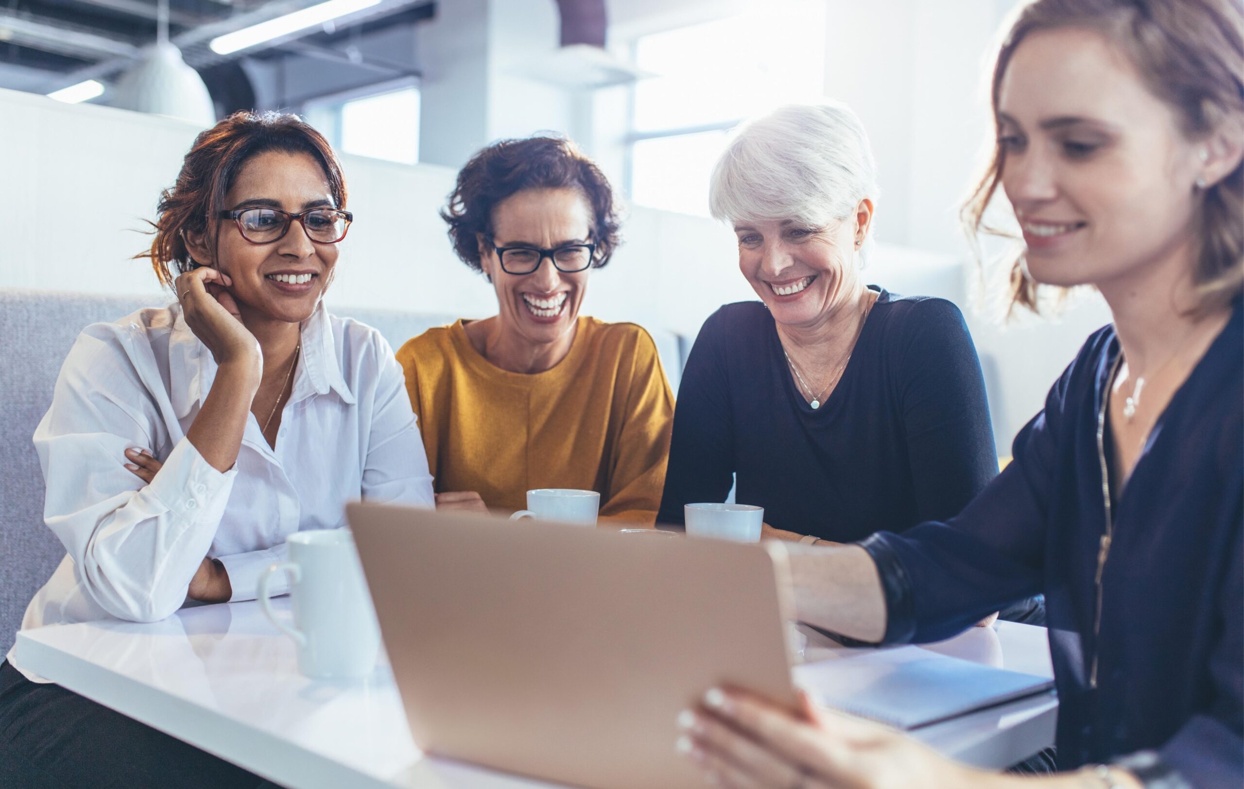 vier Frauen mit einem Kaffee schauen sich etwas auf dem Laptop an und lachen