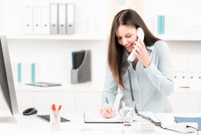 Frau hat lächelnd Telefon in der Hand und notiert sich etwas auf einem Block